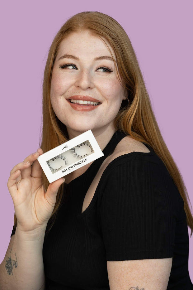 Redhead model holding up a box of fake eyelashes close to her face. The model is smiling and looking to the side.